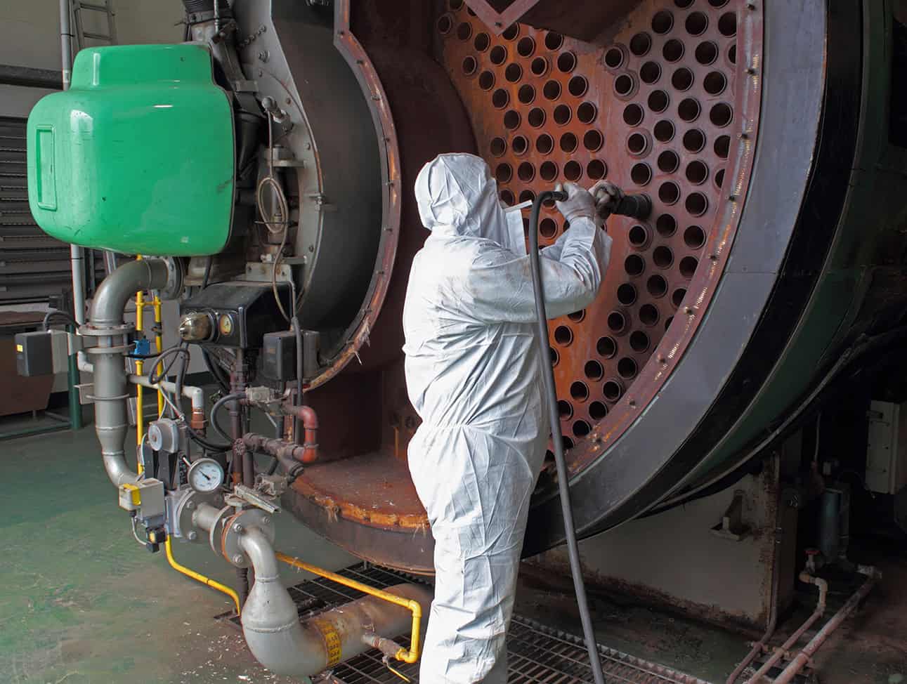 A boiler technician wearing protective gear cleaning a large boiler
