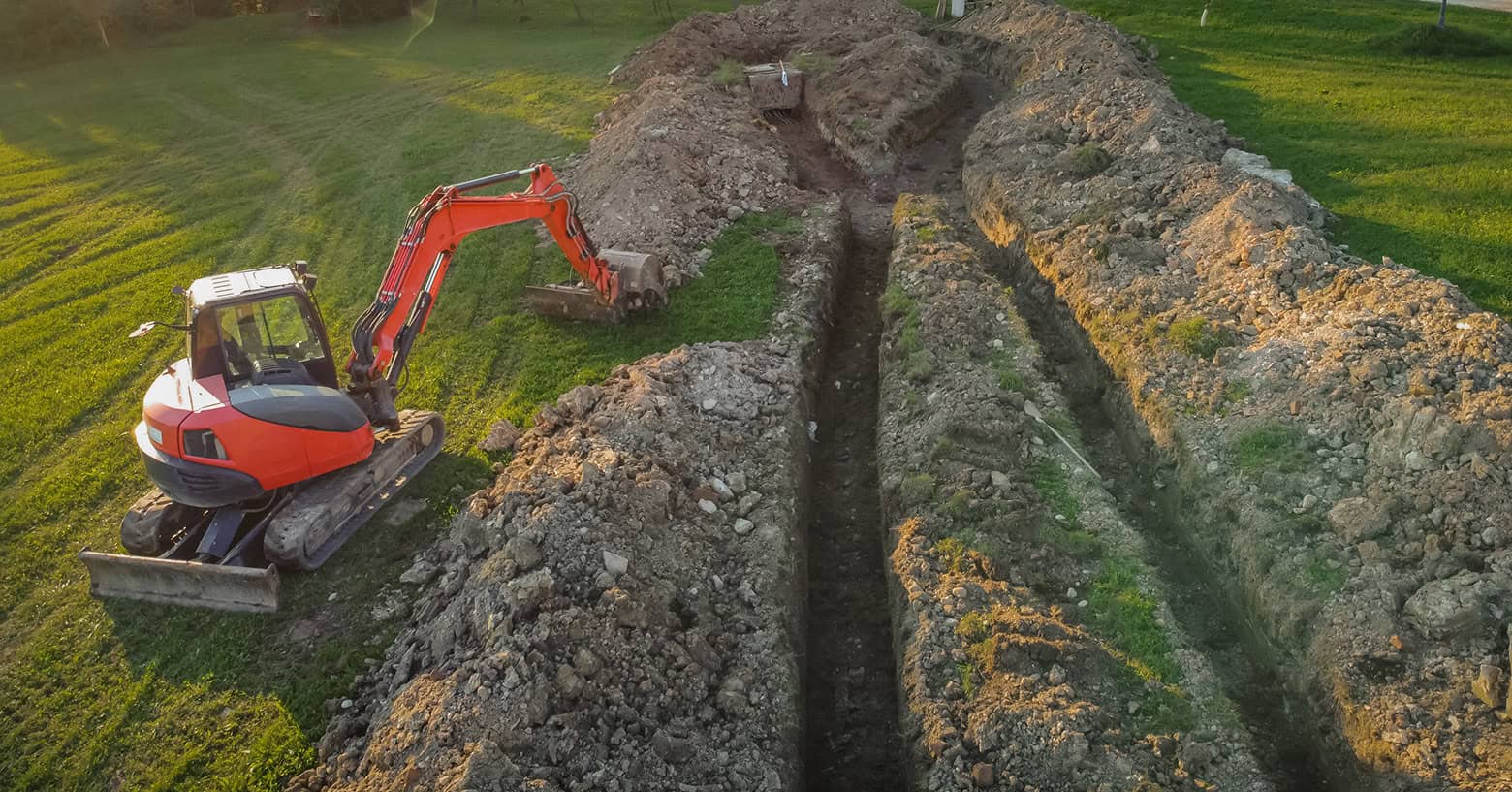 An excavator digging a shallow trench across a long field for a horizontal ground loop geothermal heat pump system.